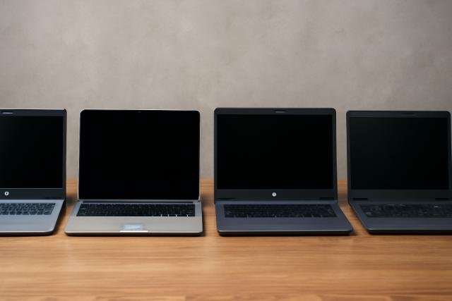 A row of four laptops (Dell, Apple, HP, and Lenovo) with black screens, placed on a wooden surface against a soft gray concrete background.